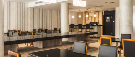 A modern dining area with a long communal table and individual tables, surrounded by chairs with wooden frames and black cushions.