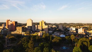 Une vue urbaine avec différents bâtiments, dont un imposant bâtiment rouge et une haute structure blanche. On aperçoit un parc avec des arbres et une fontaine au premier plan.