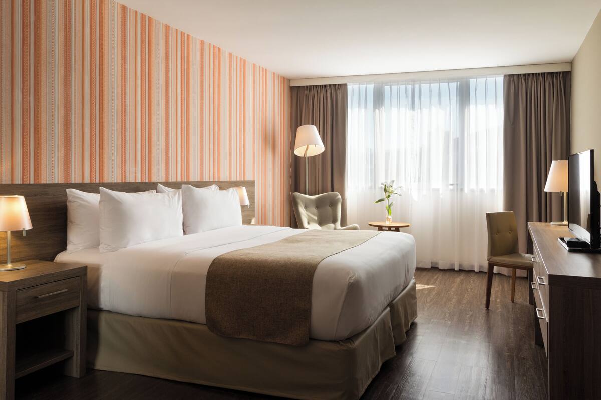A hotel room with a large bed, two bedside tables with lamps, a desk with a chair, a television, and a window with curtains.