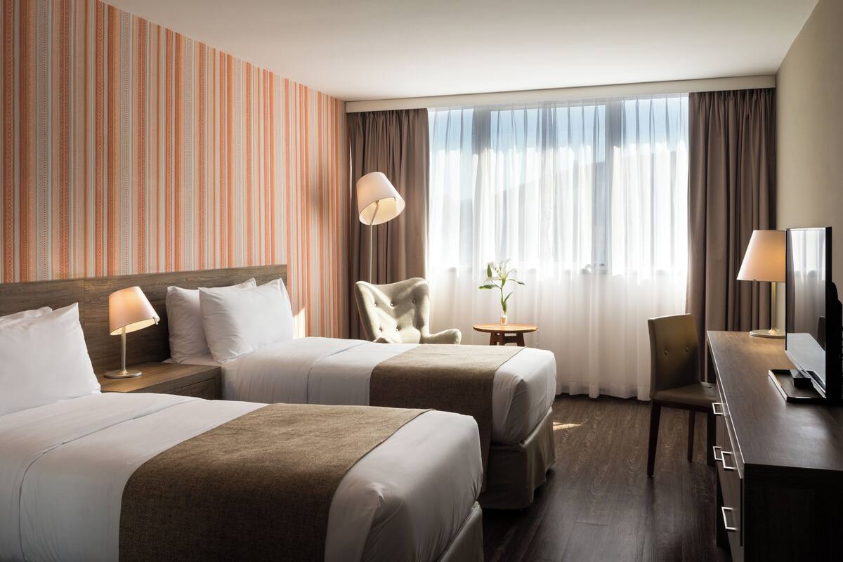 A hotel room with two beds, a desk, a chair, a lamp, and a view of the outdoors.