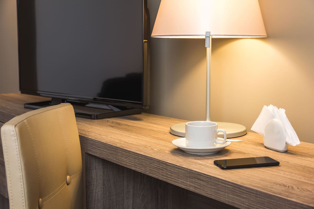 A hotel room with a wooden desk, a television, a white cup and saucer, a smartphone, and a lamp.