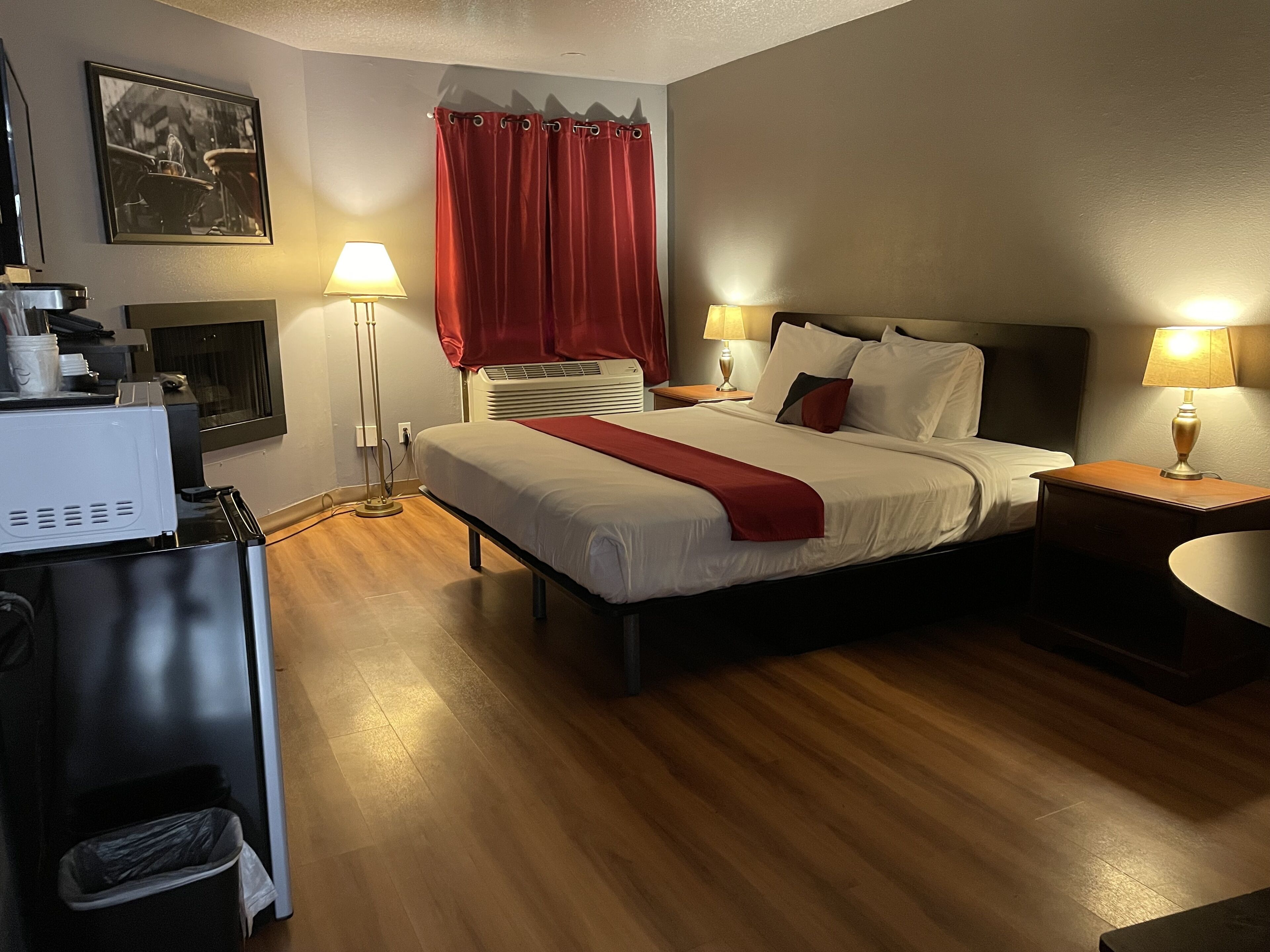 Habitación de hotel con cama, dos mesitas de noche, lámpara, calefactor, un cuadro en la pared y ventana con cortinas rojas.