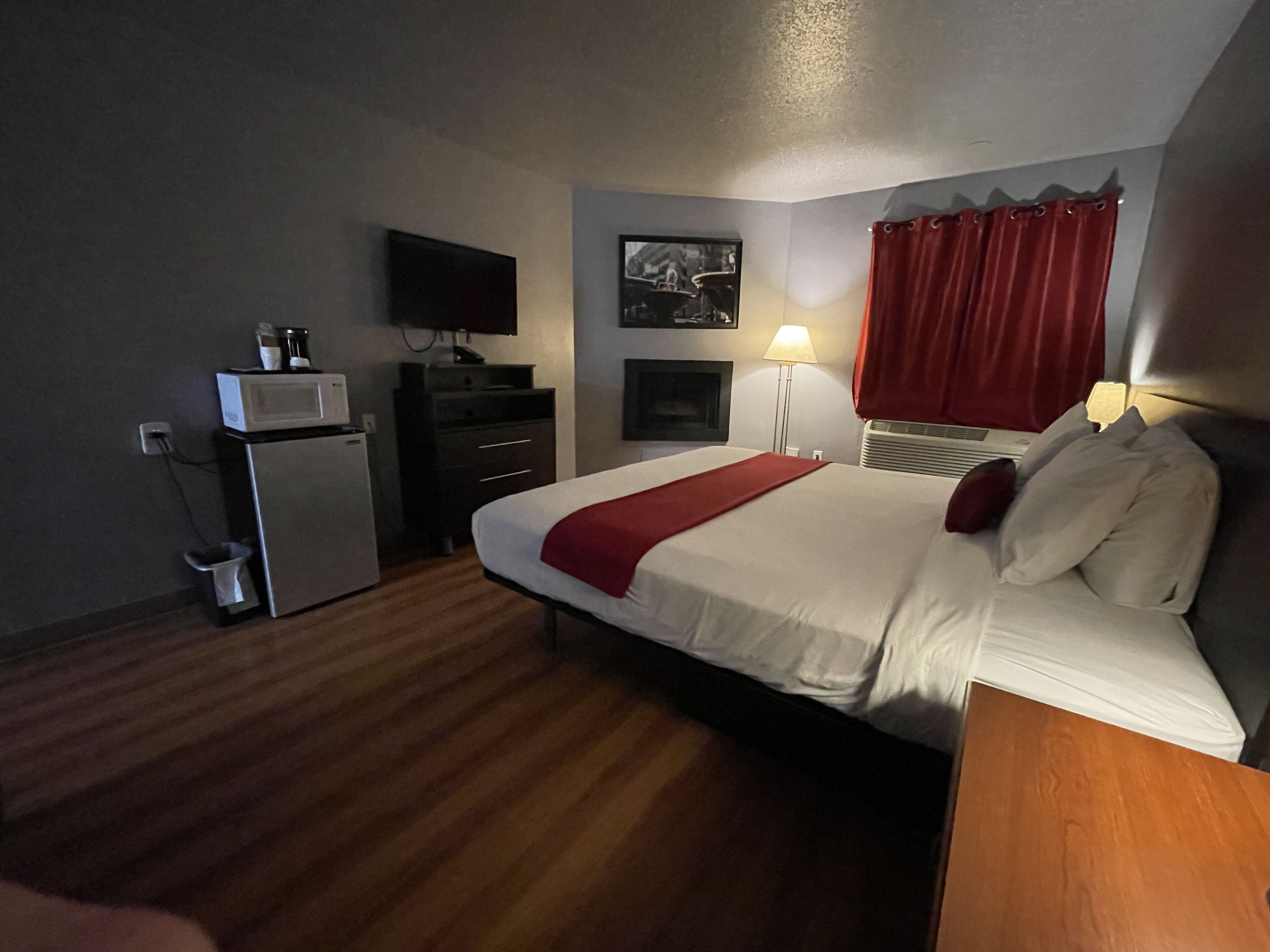 A hotel room with a bed, a desk, a microwave, a TV, a lamp, and a red curtain.