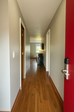 A hallway with wooden flooring, a red wall, and a door with a modern handle. - Sunnyside Inn & Suites (Clackamas)