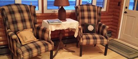 L’intérieur chaleureux d’un chalet avec deux fauteuils à motifs écossais, une petite table avec une lampe et une vue sur un paysage enneigé par les fenêtres.