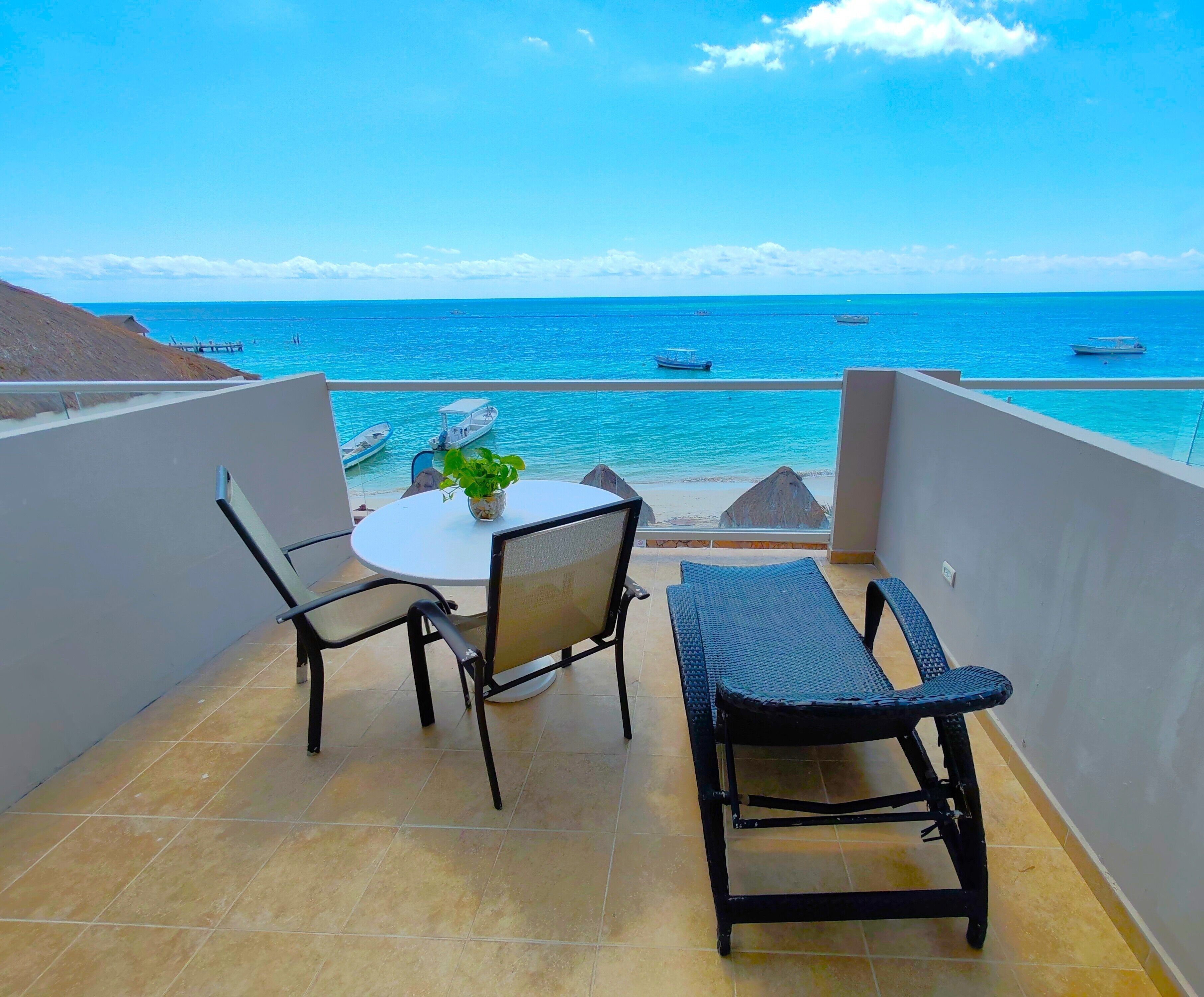 Uma varanda com mesa e cadeiras, um vaso de planta e vista para o mar com barcos.