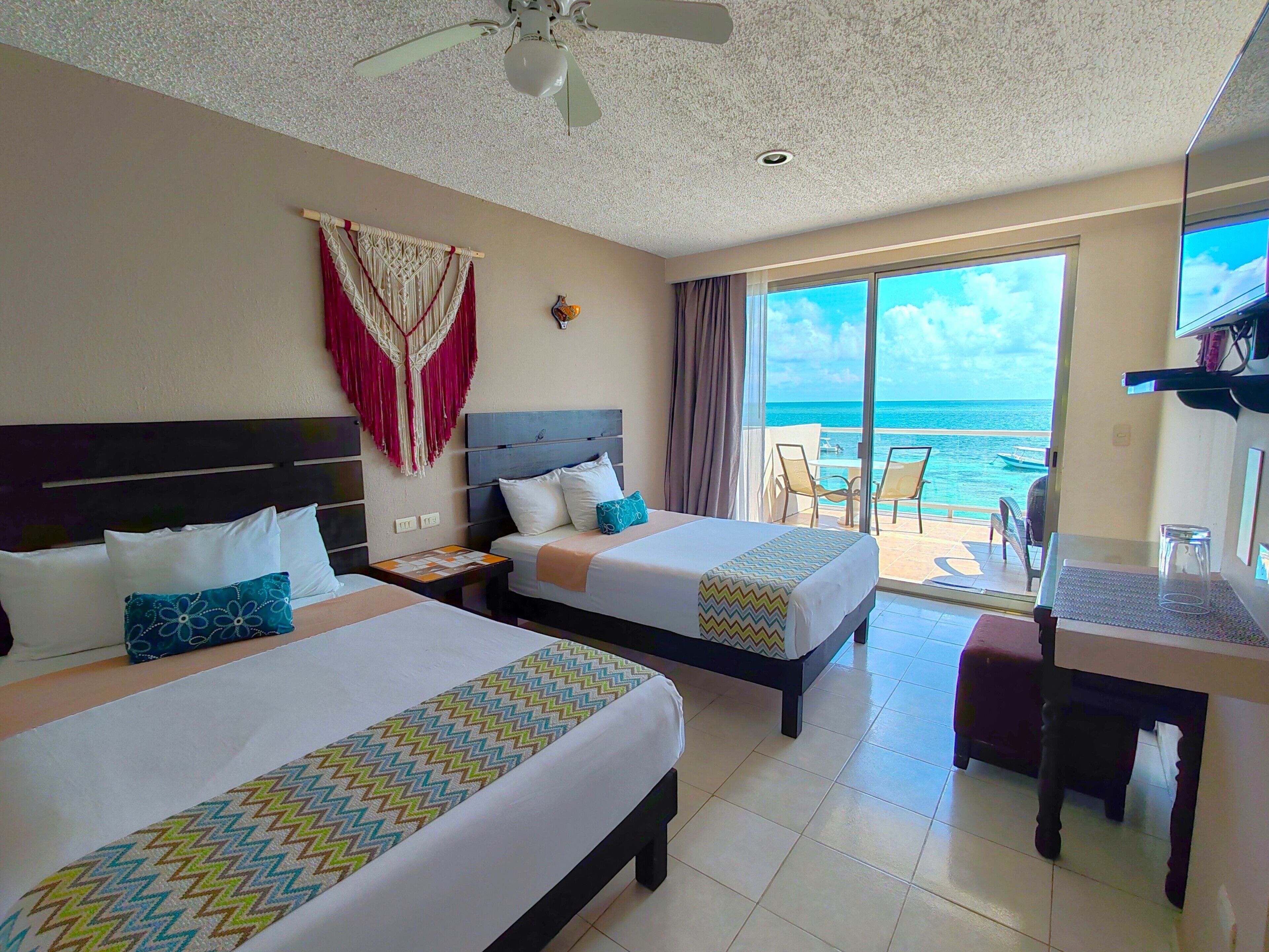 A hotel room with two beds, a ceiling fan, a large window with a view of the ocean, and a table with chairs.