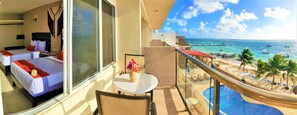 Two beds with red pillows and white bedspreads, a small table with a vase of flowers, and a balcony with a view of the beachfront resort.