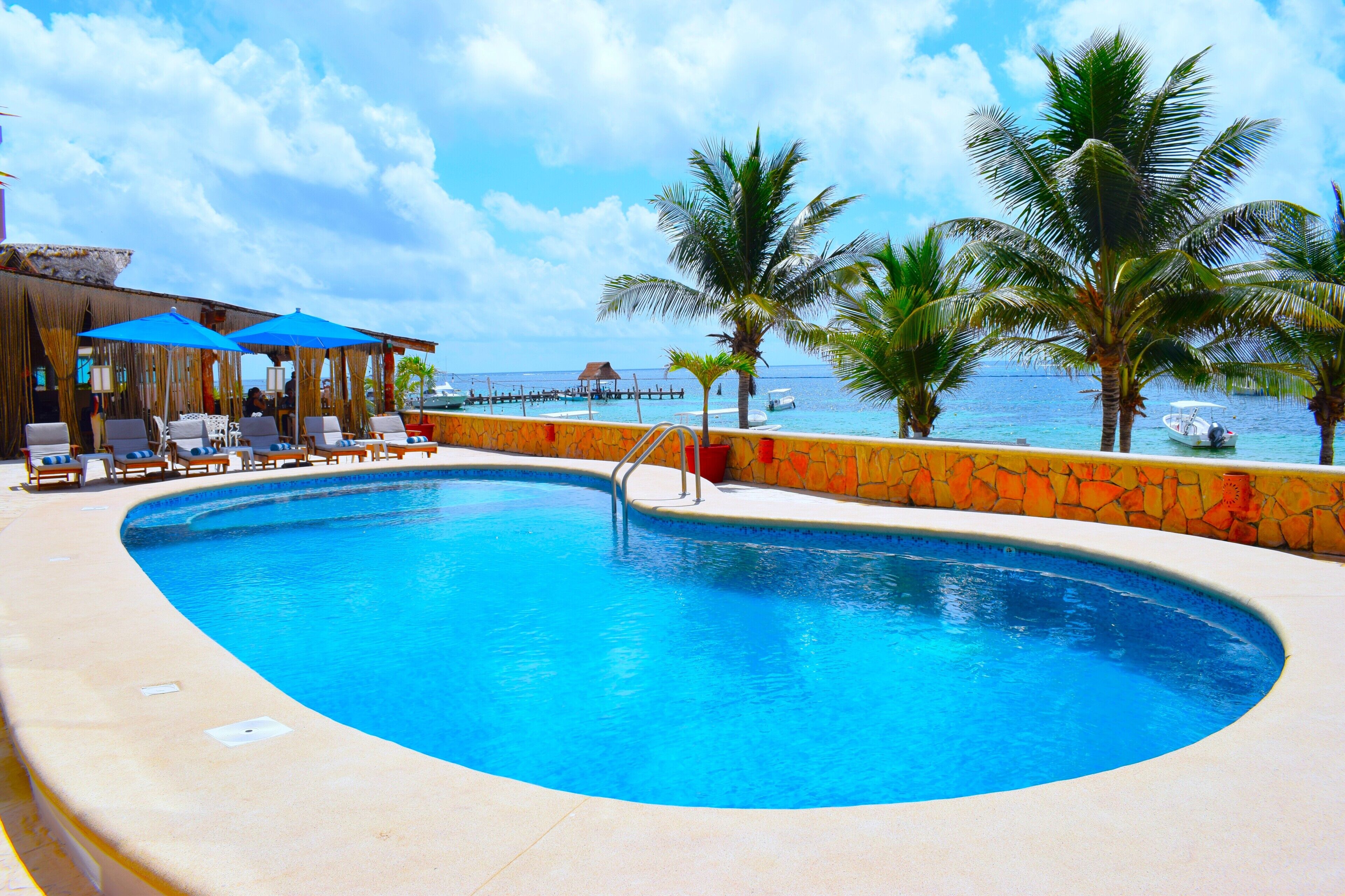 A pool with lounge chairs and umbrellas, a stone wall, and a view of the ocean.