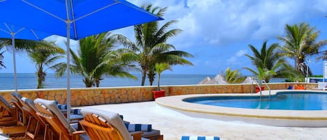 Ein Poolbereich mit Liegestühlen, einem blauen Sonnenschirm und Blick auf das Meer.