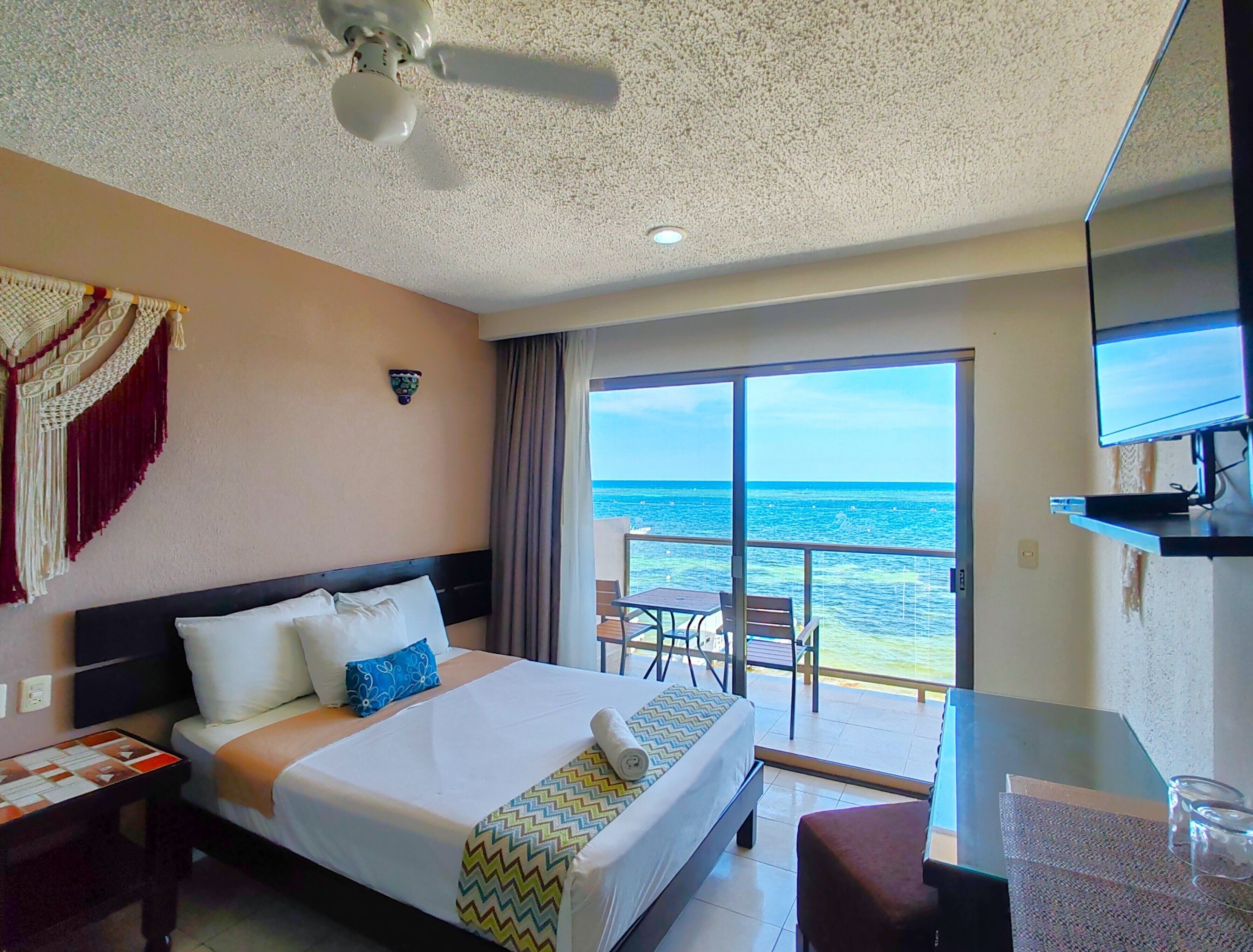 Habitación de hotel con cama, ventilador de techo, televisión, balcón con vista al mar y una mesa.
