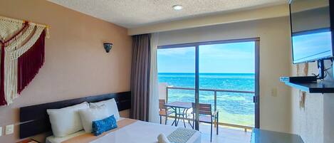 Habitación de hotel con cama, ventilador de techo, televisión, balcón con vista al mar y una mesa.