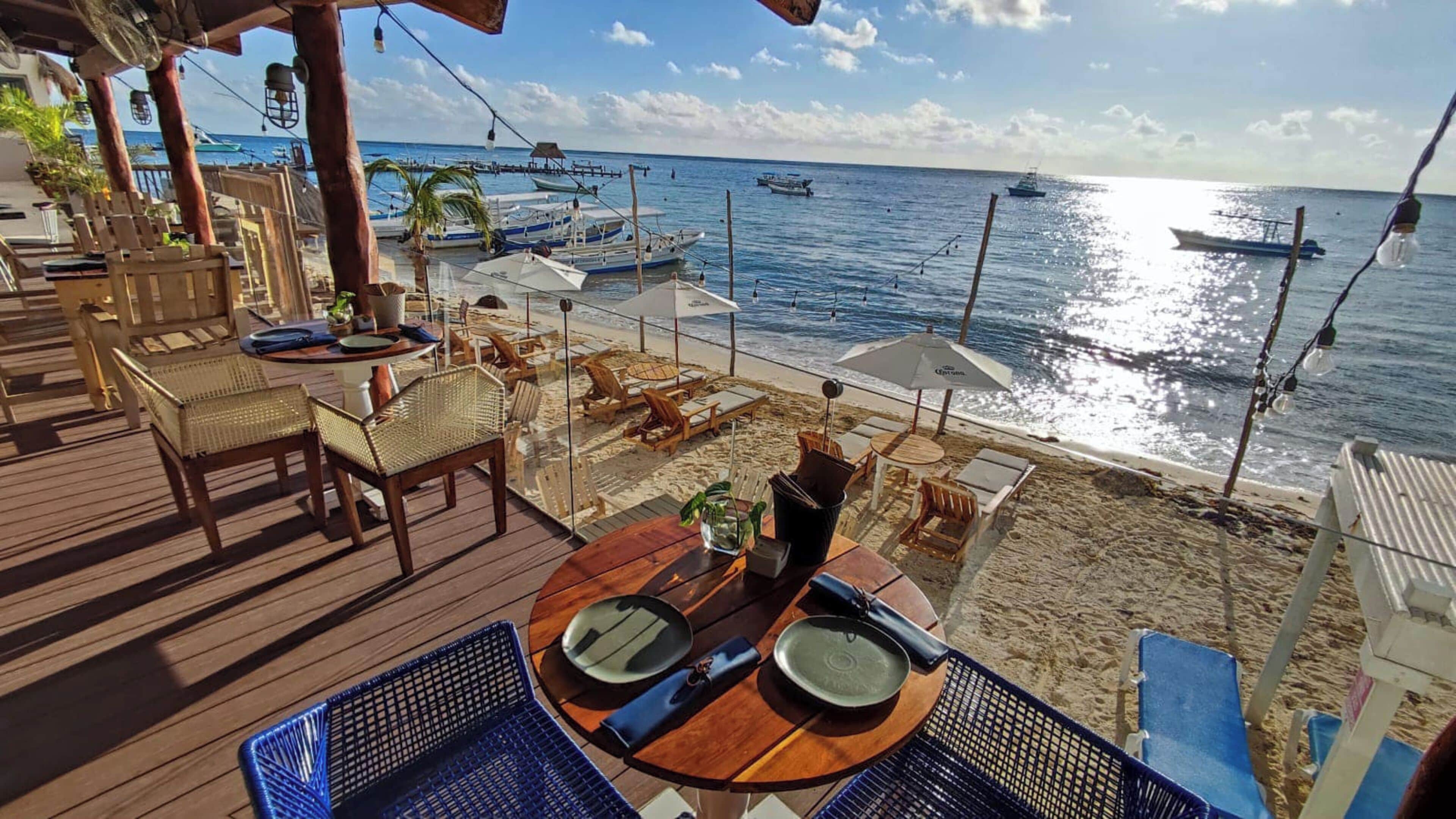 A beachside restaurant with wooden deck seating, tables set for dining, and a view of the ocean with boats.