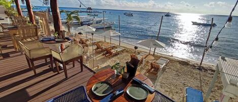 A beachside restaurant with wooden deck seating, tables set for dining, and a view of the ocean with boats.