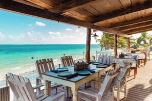 An outdoor dining area with a wooden deck overlooking the ocean, featuring a table set for two with chairs. - Hacienda Morelos Beach Front Hotel (Puerto Morelos)