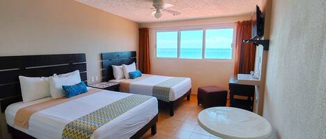 Une chambre d’hôtel avec deux lits, un ventilateur de plafond, une petite table et une vue sur la mer.