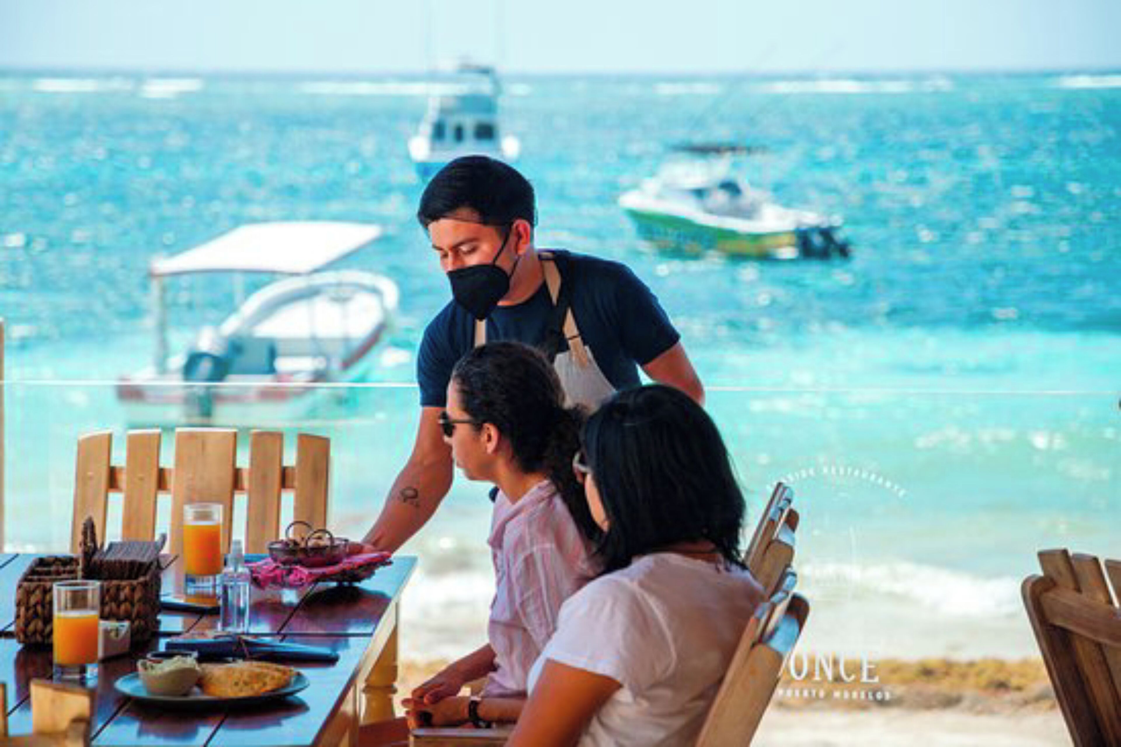 Une table à manger avec de la nourriture et des boissons, une personne qui sert, et une vue sur la mer et les bateaux.