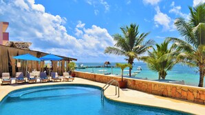 Ein Poolbereich mit Liegestühlen und Sonnenschirmen mit Blick auf einen Strand, gesäumt von Booten und Palmen.