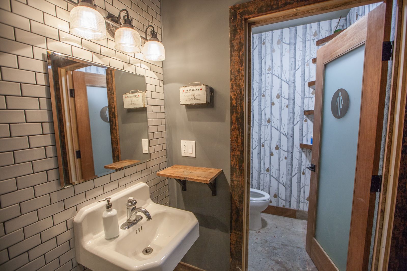 A bathroom with a white sink, mirror, and wooden shelf. There is a First Aid Kit and a toilet visible.