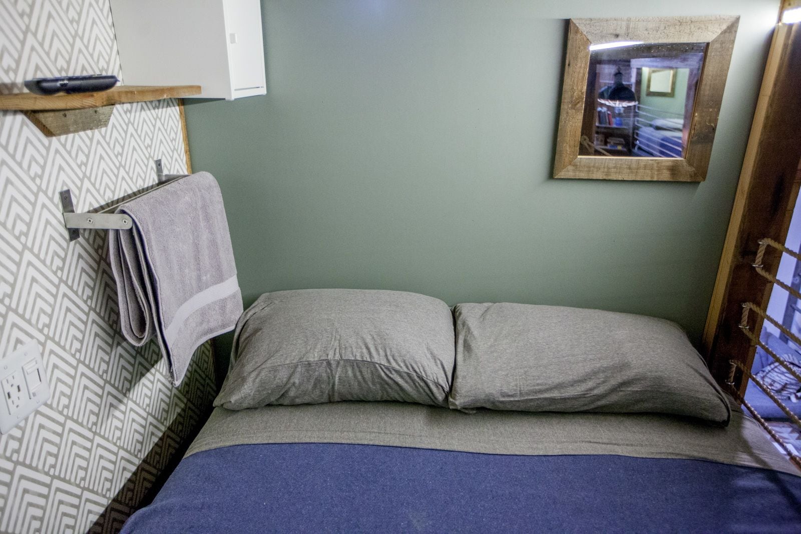 A small, single-bed room with a grey wall, a wooden shelf with a towel, and a framed mirror on the wall.