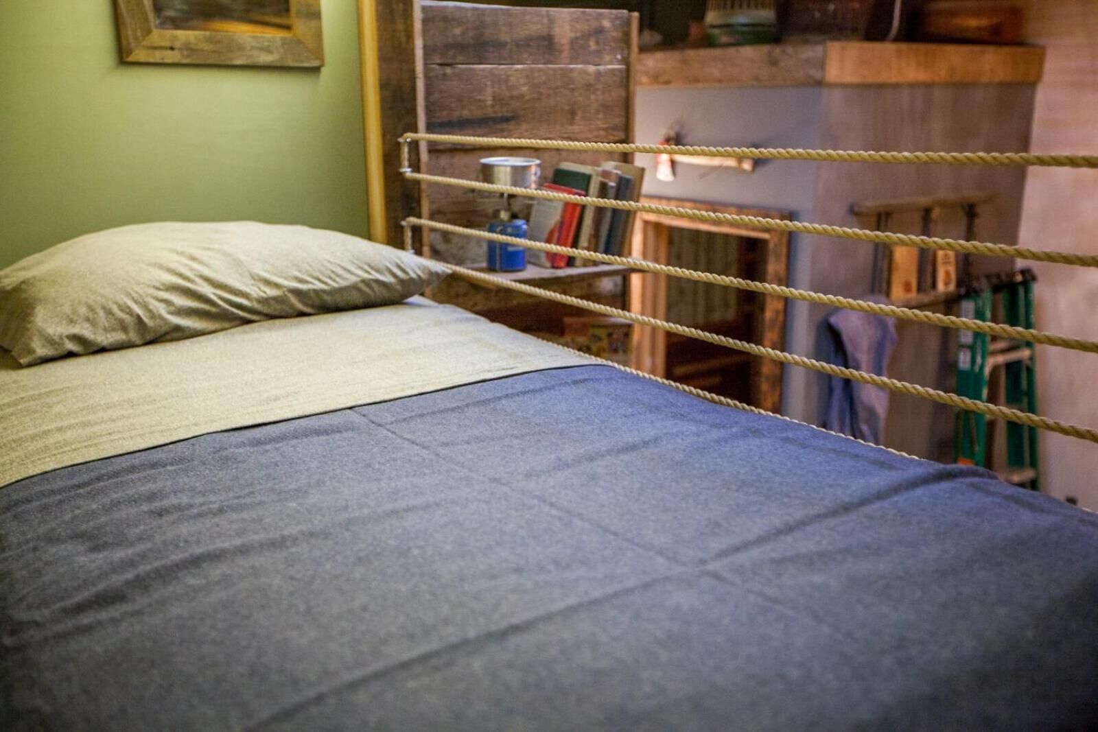 A bed with a blue blanket and a pillow in a room with a wooden bunk bed and a rope barrier.