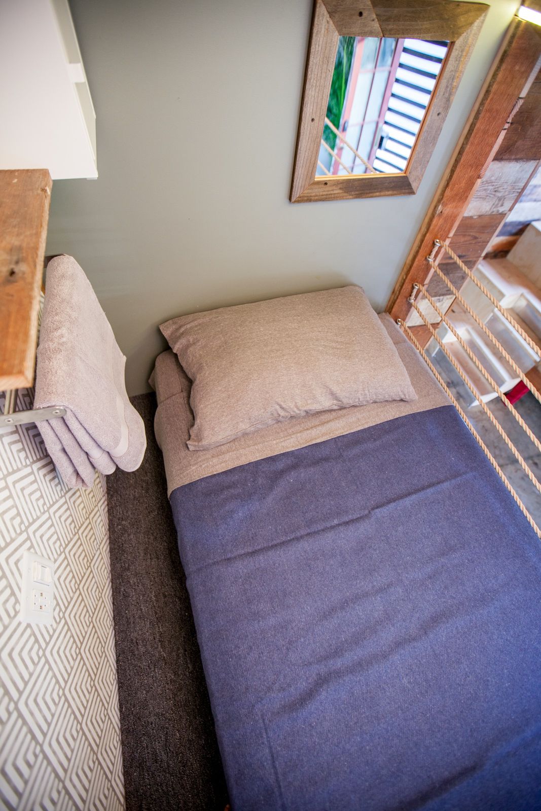 Un petit lit bien fait, avec une couverture bleue et deux oreillers, une tête de lit en bois, une petite table en bois sur laquelle est posée une serviette, et une fenêtre donnant sur des immeubles.