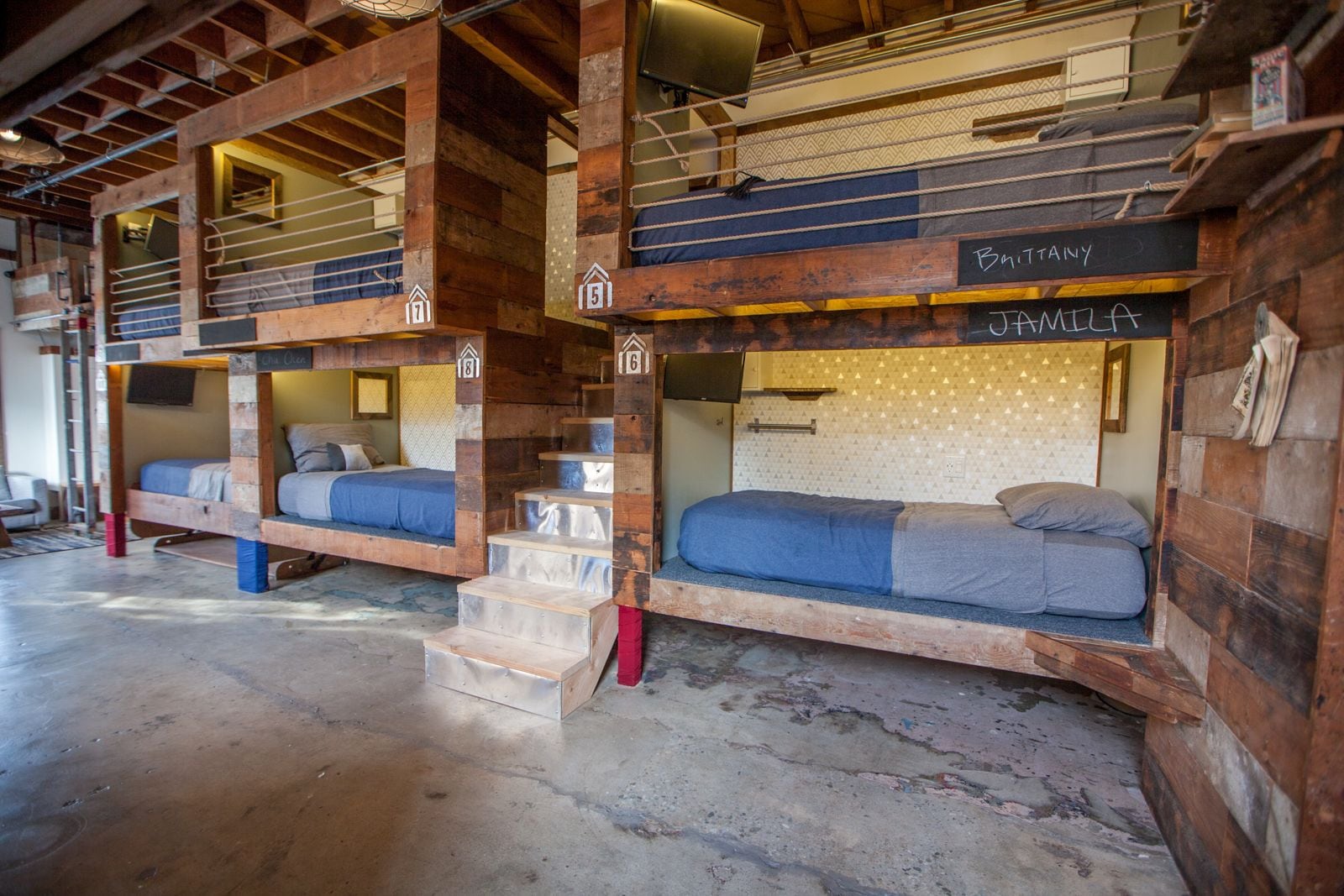 A rustic cabin-style hotel with bunk beds labeled "Brittany" and "Familia," wooden beams, and a concrete floor.