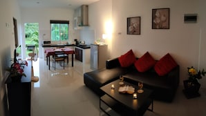 Een moderne woonkamer met een zwarte bank, rode kussens en een salontafel met drankjes en een boek. Op de achtergrond is een eethoek met een tafel en stoelen zichtbaar.