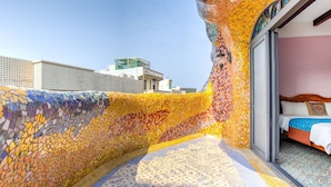 A room with a mosaic wall, a bed with patterned bedding, and a view of the cityscape.