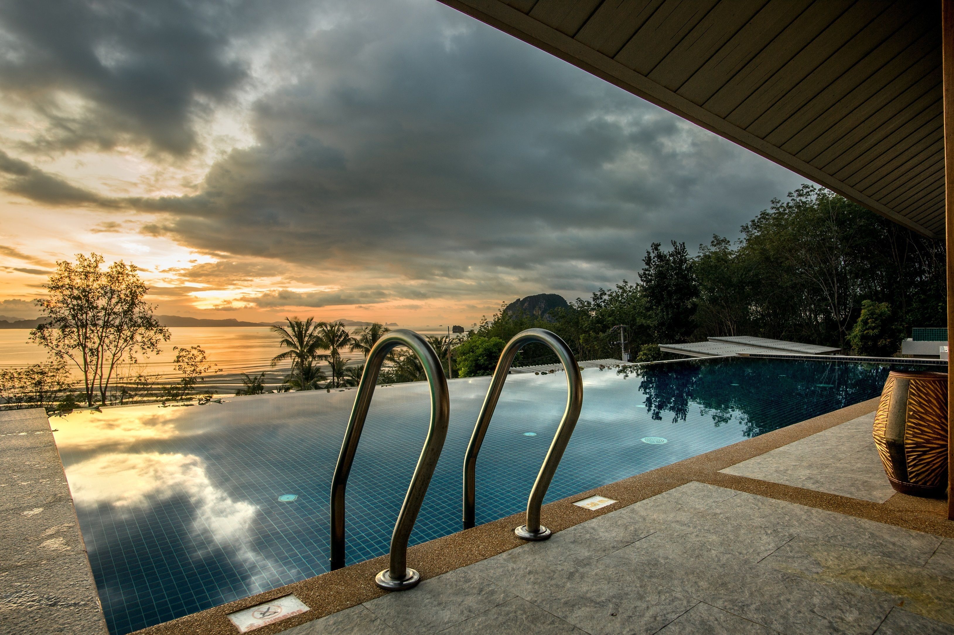 Uma piscina infinita com grades de metal, vista do pôr do sol sobre o oceano e paisagem tropical com palmeiras.