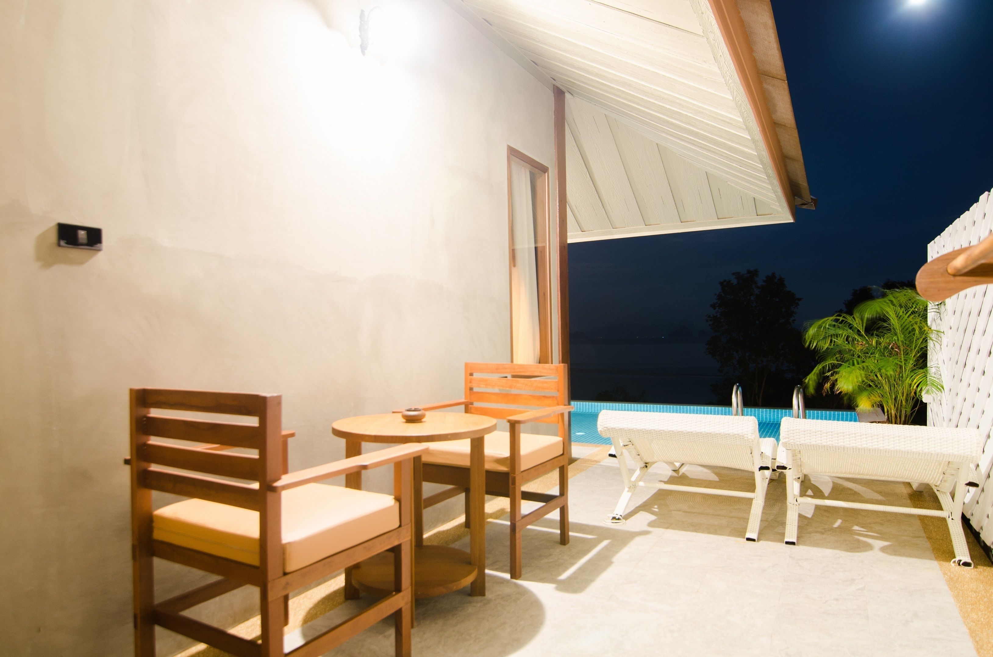 A balcony with wooden chairs and a small table, overlooking a swimming pool and palm trees.