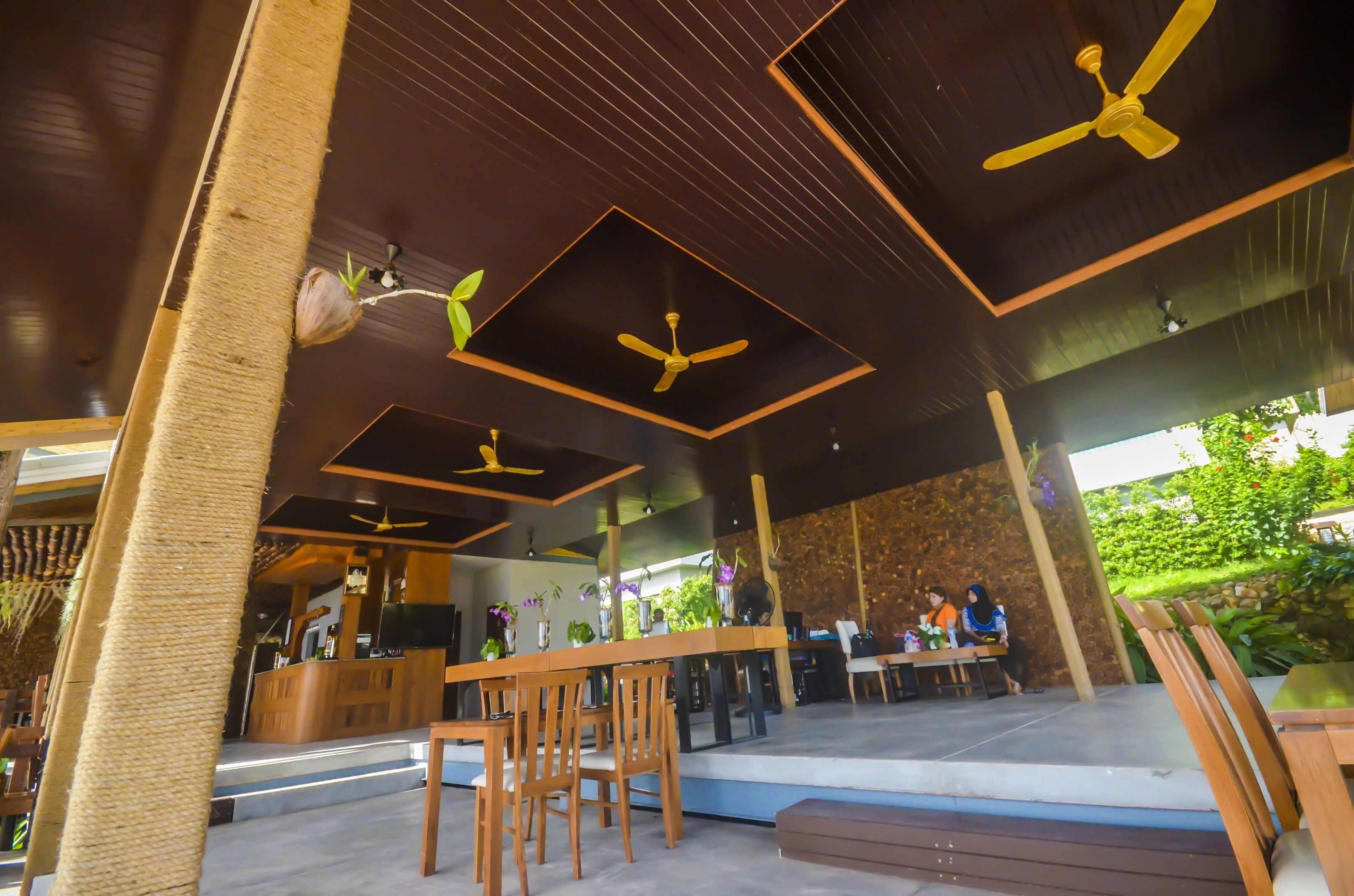 A modern outdoor seating area with wooden tables and chairs, ceiling fans, and a reception desk.