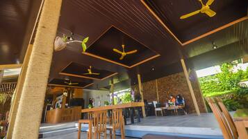 A modern outdoor seating area with wooden tables and chairs, ceiling fans, and a reception desk.