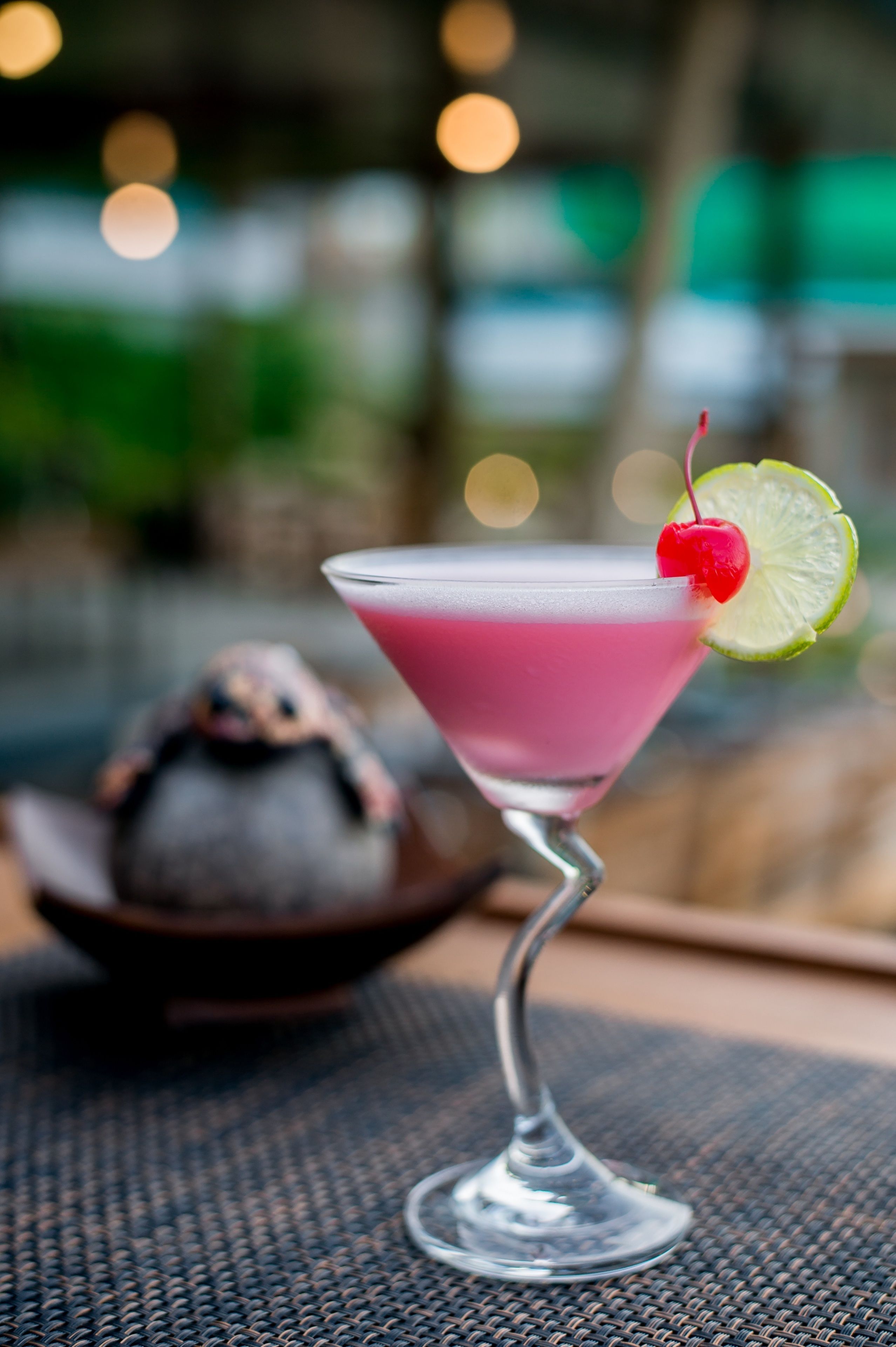 A pink cocktail with a cherry and lime garnish on a martini glass, placed on a woven table mat.