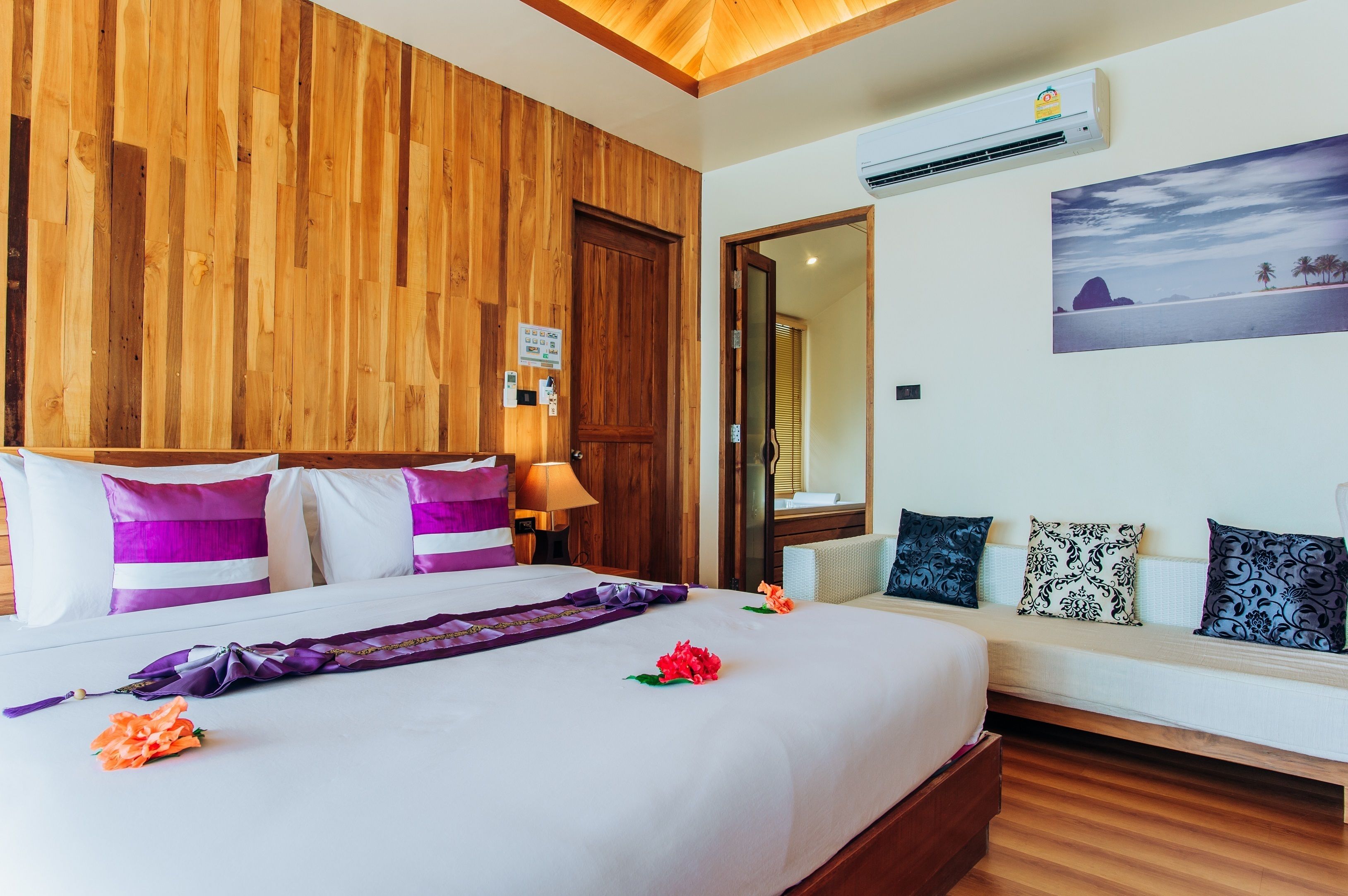 Ein Hotelzimmer mit einer Wand aus Holzpaneelen, einem Bett mit violett-weißen Streifen-Kissen, einem weißen Sofa mit gemusterten Kissen und einem Wandbild mit einer Strandansicht.