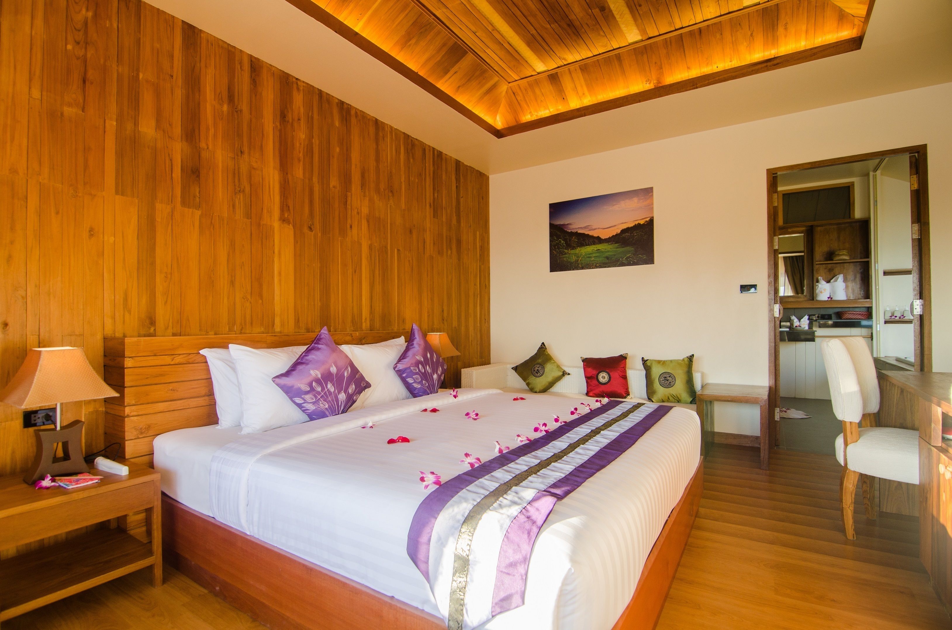 Habitación de hotel con techo de madera, una cama grande con colcha blanca y morada, y un cuadro en la pared.
