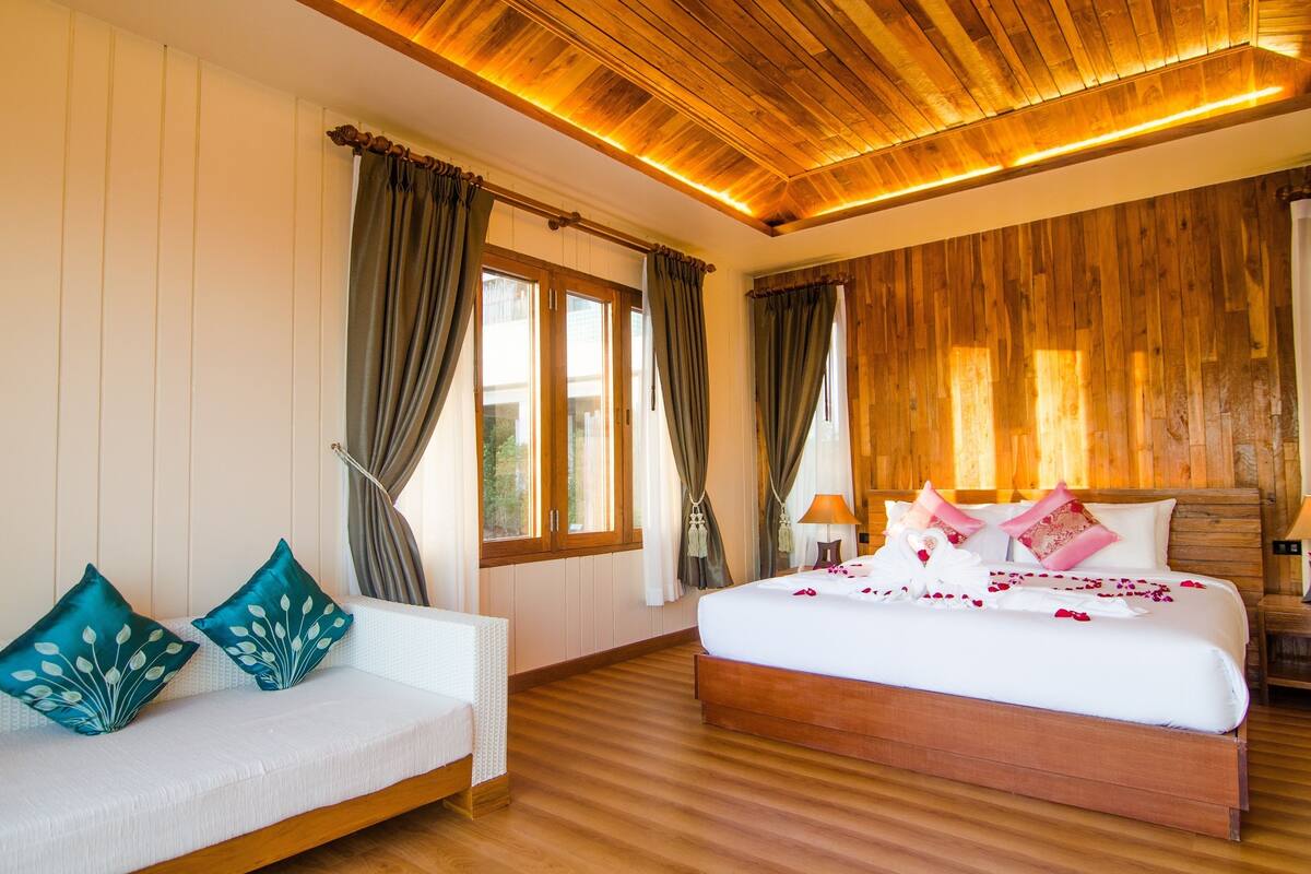 Une chambre d’hôtel avec un plafond en bois, un grand lit avec une literie blanche et des oreillers roses, un canapé blanc avec des coussins turquoise et un plancher en bois.