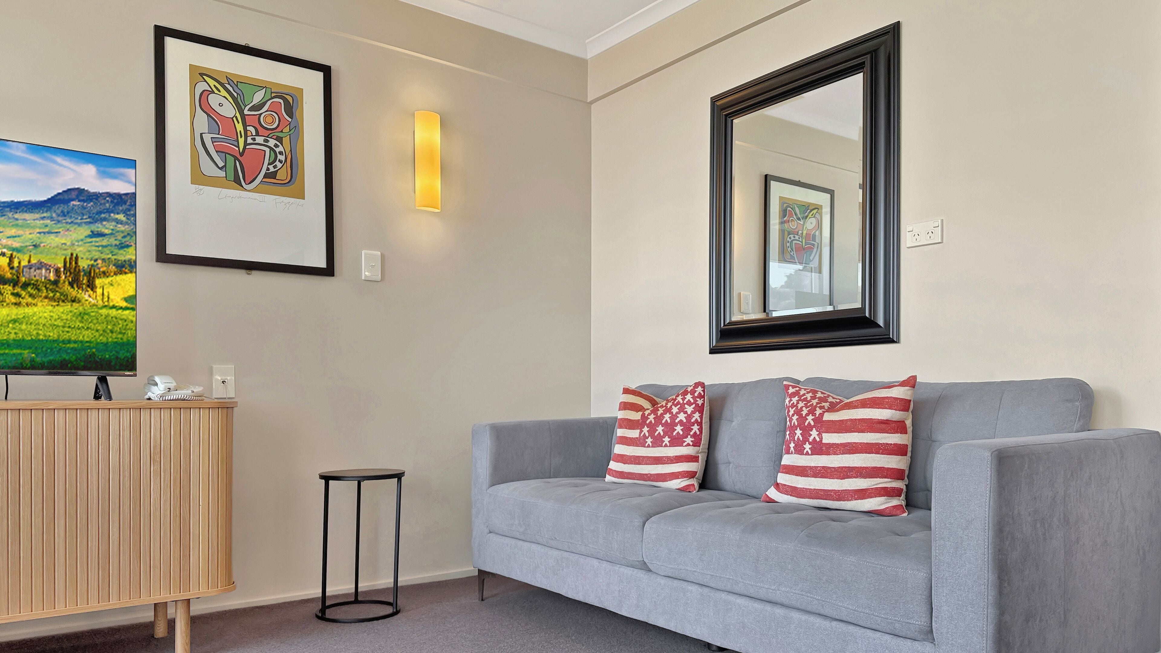 A modern living room with a grey sofa, two American flag pillows, a framed abstract painting, a flat-screen TV, and a wooden side table.