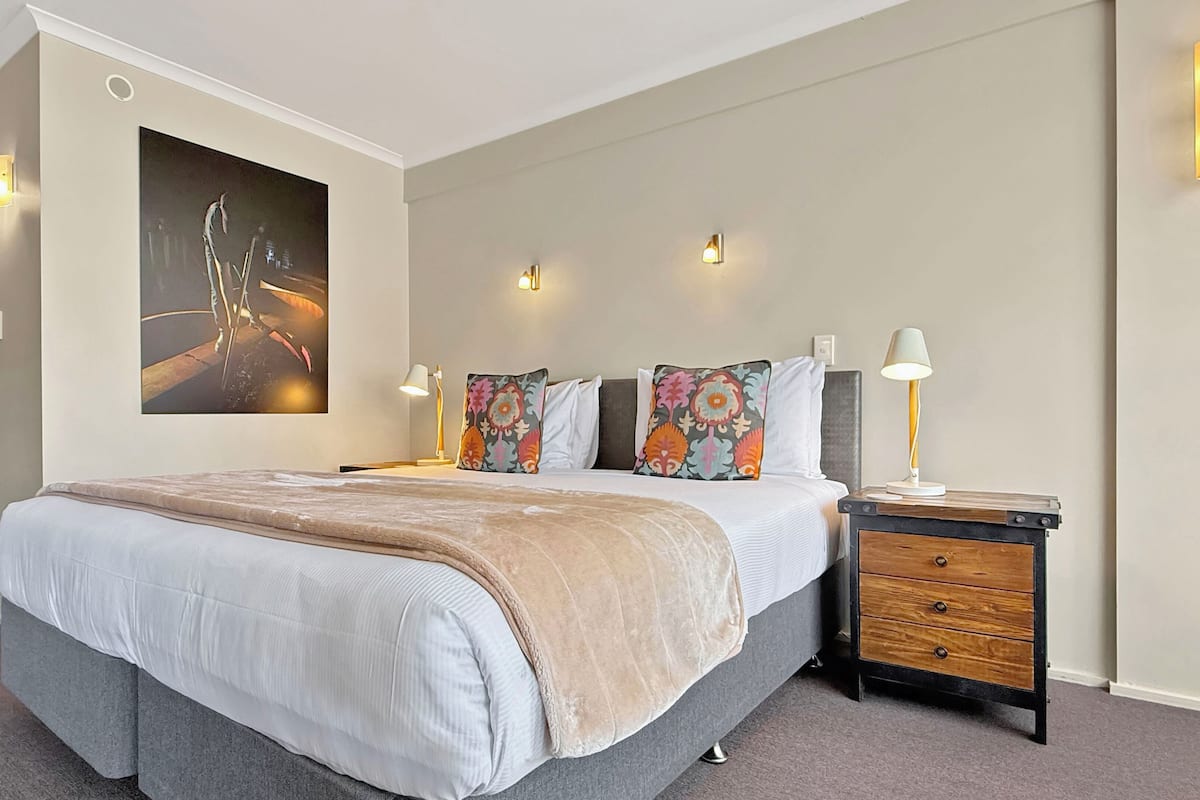 A modern bedroom with a large bed, decorative pillows, a bedside table with a lamp, and a wall-mounted light fixture.