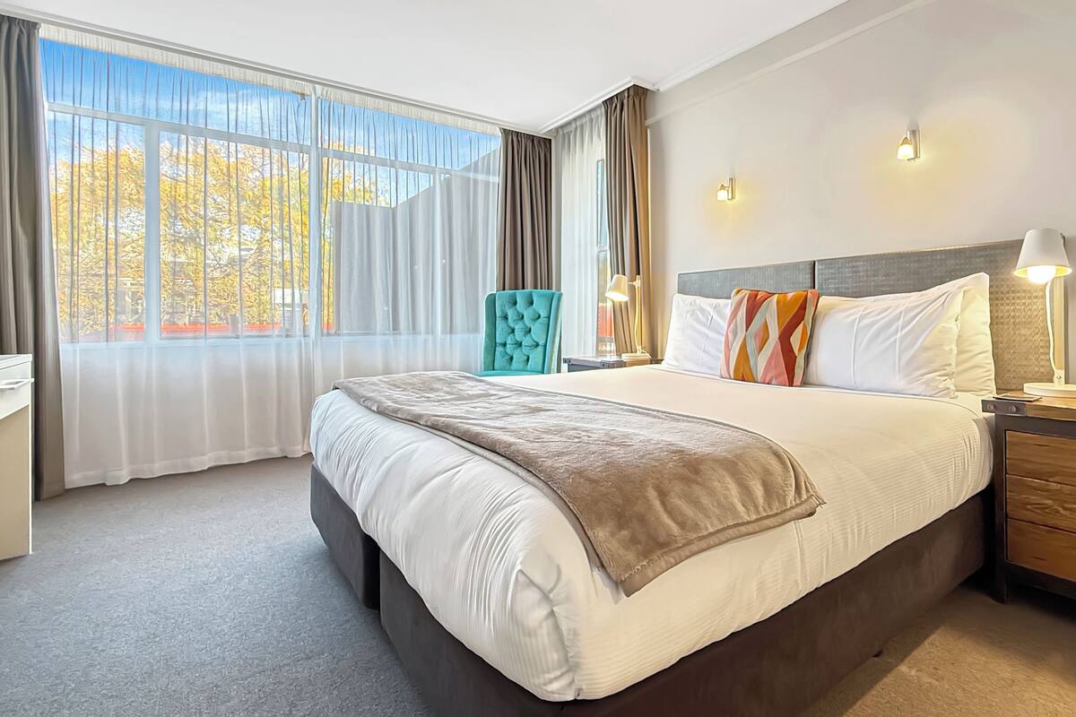 A hotel room with a large bed, a desk, a chair, and a nightstand with a lamp.