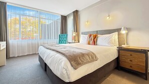 A hotel room with a large bed, a desk, a chair, and a nightstand with a lamp.
