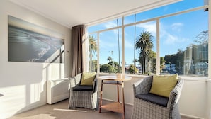 A room with two wicker chairs, a small round table, a large window with a view of palm trees and a cityscape, and a framed picture on the wall.