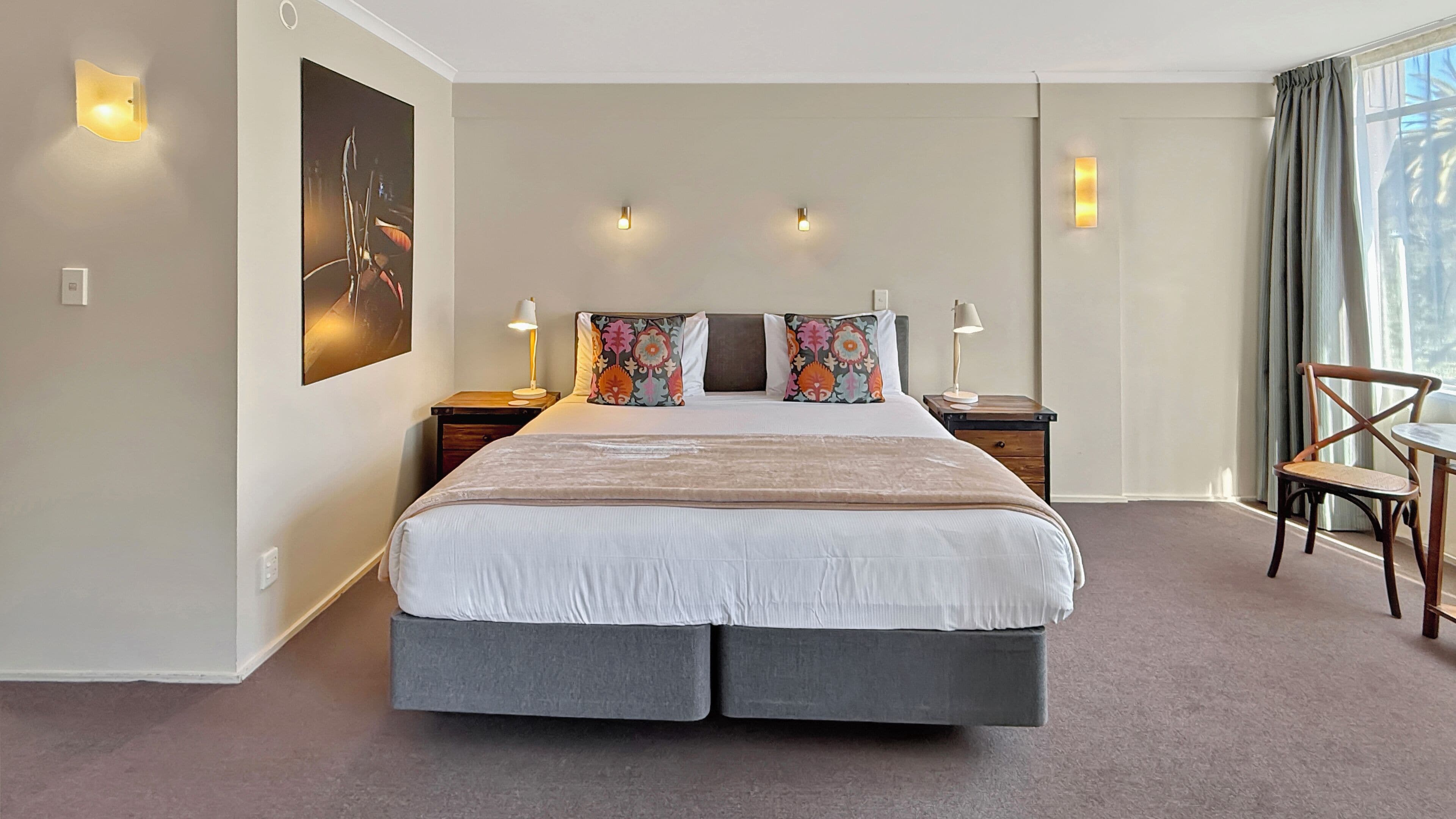 A hotel room with a large bed, two bedside tables with lamps, a chair, and a large window with curtains.