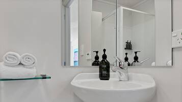 Un baño con un lavamanos blanco, dos dispensadores de jabón negros y un espejo que refleja un perchero con toallas enrolladas.