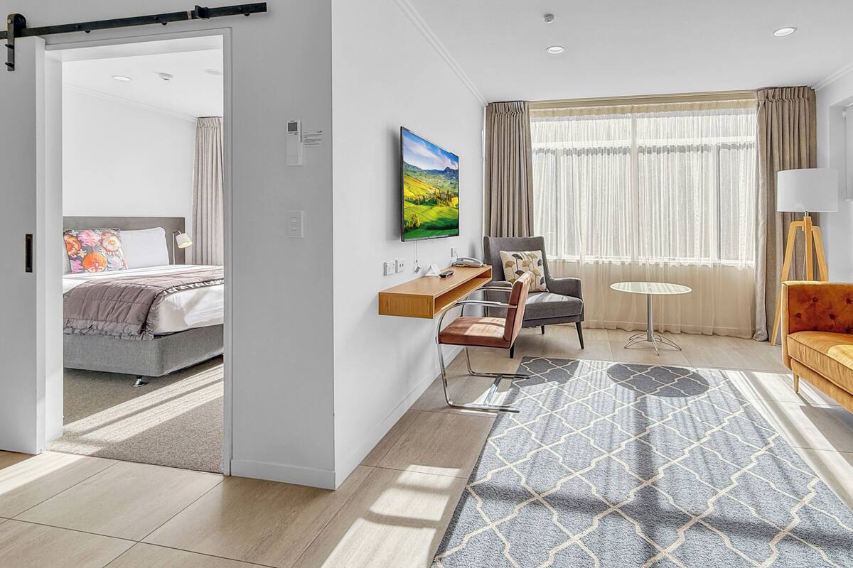 A hotel room with a bed, a desk, a chair, a sofa, a TV, and a window with curtains.