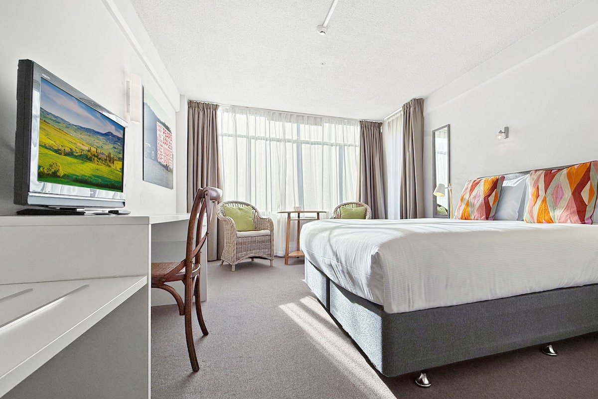 A hotel room with a bed, a desk with a television, a chair, a small table, and a window with curtains.