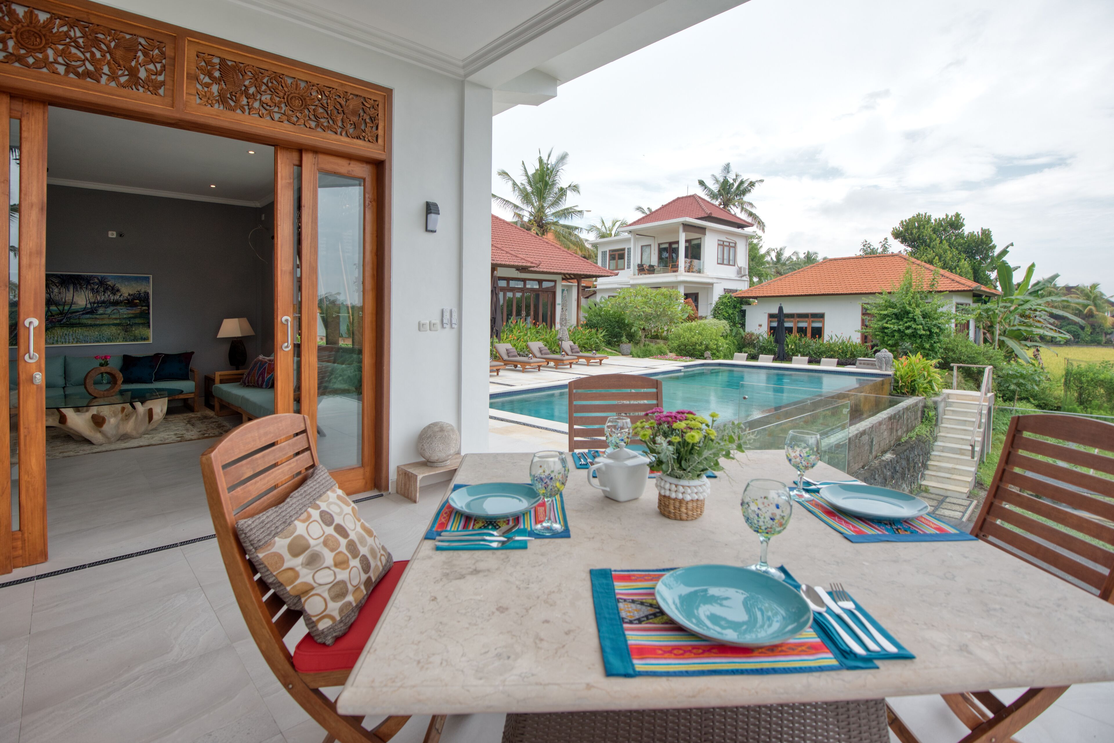 A dining area with a table set for four, a pool in the background, and a view of a house with a red roof.