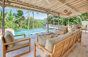Un área de estar al aire libre con techo, piscina, muebles de madera y exuberante vegetación al fondo.