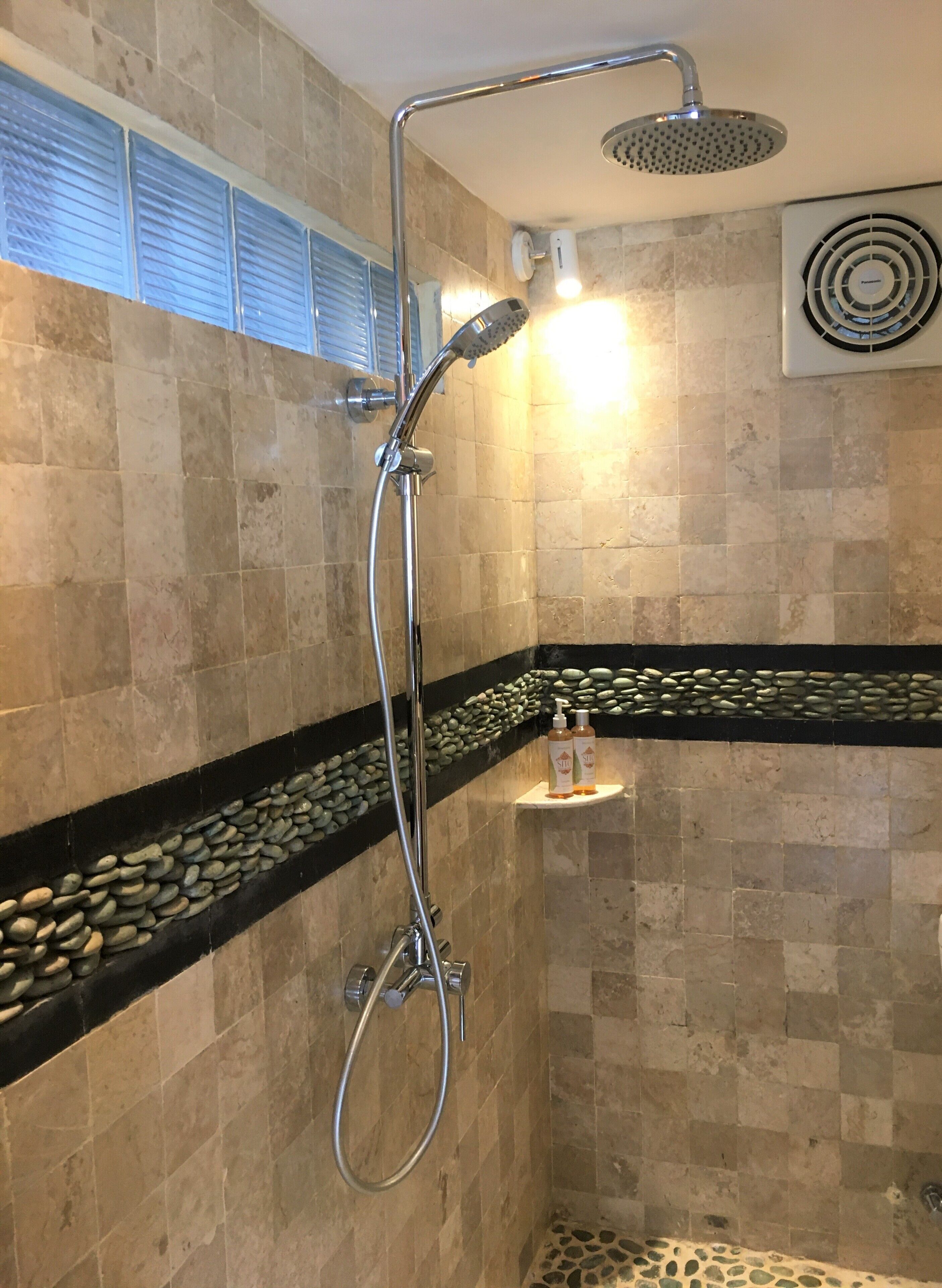 A shower with a handheld showerhead and a wall-mounted rain showerhead, a shelf with toiletries, and a decorative stone border.