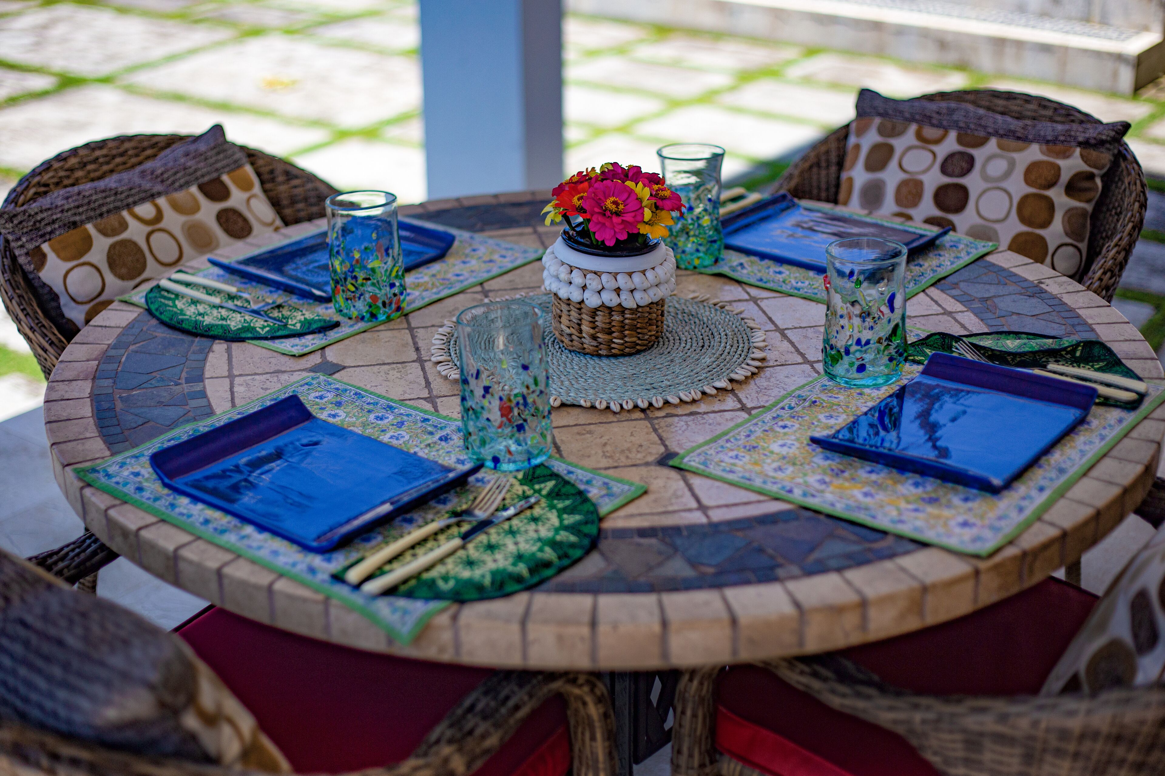 Una mesa redonda para comer al aire libre con un centro tejido, individuales azules y verdes, y sillas de mimbre con cojines estampados.
